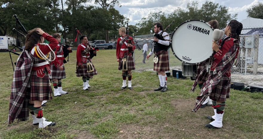 Eight bagpipers and drummers in Scottish kilts stand in a semi circle, warming up on a grass field
