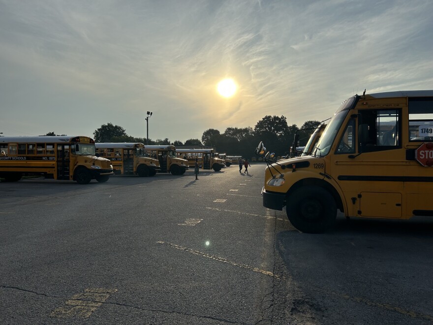 Buses are parked in a parking lot. A few people mill in between. A hazy sunrise in the background.