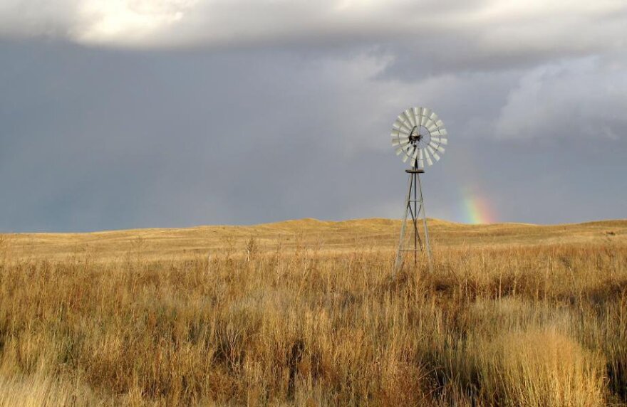 An old metal windmill stands in a field of brown grass. The sky is cloudy and there is a slight rainbow in it.