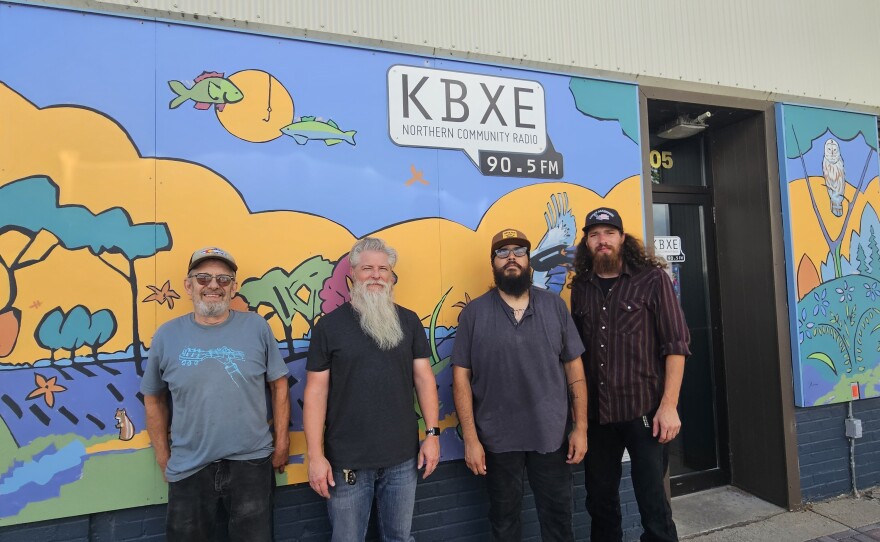 Four men standing in front of a yellow, blue, and green mural. A speech bubble over their head says "KBXE: Northern Community Radio 90.5FM"