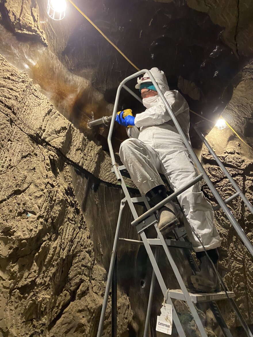 A person in a full-body, white suit with their face covered and protective glasses uses a drill-like piece of equipment to drill into a frozen dirt wall.