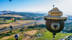 A photo taken from a hot air balloon in the sky of another hot air balloon in the shape of a bear with a hat that reads, “Smokey.” There is a bright blue sky and other hot air balloons in the background. Grass, dirt, trees, and a long road covers the ground below.