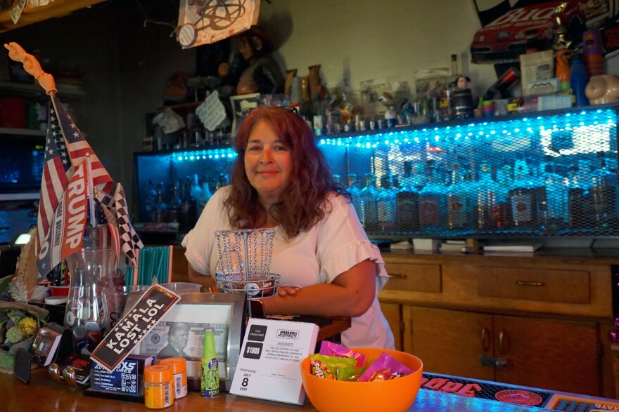 A woman with red/brown hair and a white shirt leans against a bar, with a shelf of liquor in the background and pro-Trump signs in the foreground.