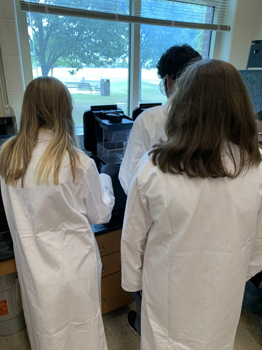 Three people are seen from the back, facing a 3D printing device that’s about the size of a typical office printer. With their fingers, they examine a tactile graphics image of a butterfly that was produced by the printer.