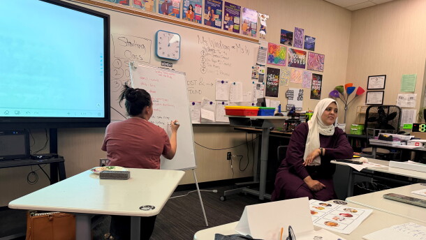 A woman on the left is writing on a whiteboard. To her right, a woman wearing a hijab talks to her class.