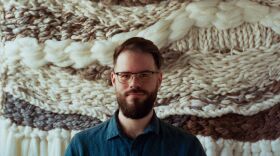 A man with glasses, a beard and blue shirt. In the background a wall hanging made of wool-like fabrics
