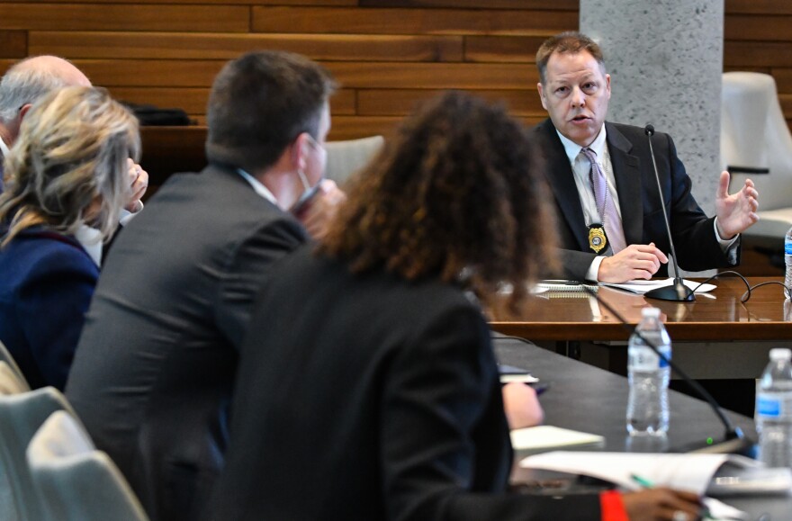 Man in suit, seated at right of a table, gestures while four people seated at left with back to camera listen.