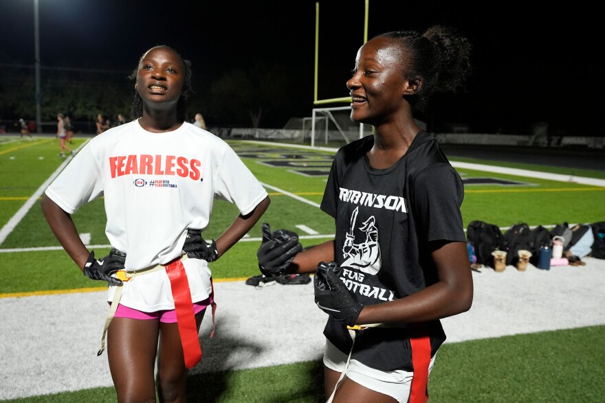 Two young girls, one wearing a white uniform outfit and the other wearing a black uniform outfit stand on a football field at night. They're both wearing belts made of red flags around their waists. 