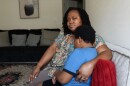 Black woman (mom) in print blouse holds a young Black boy in a light blue T-shirt, both sitting on a gray couch in a living room 