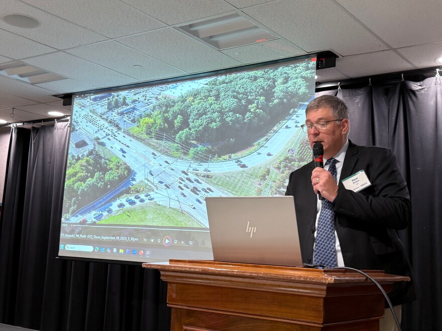 A man in a dark suit jacket and blue tie stands at a podium, reading from a laptop, with the image of a busy traffic intersection on a large multimedia screen behind him.