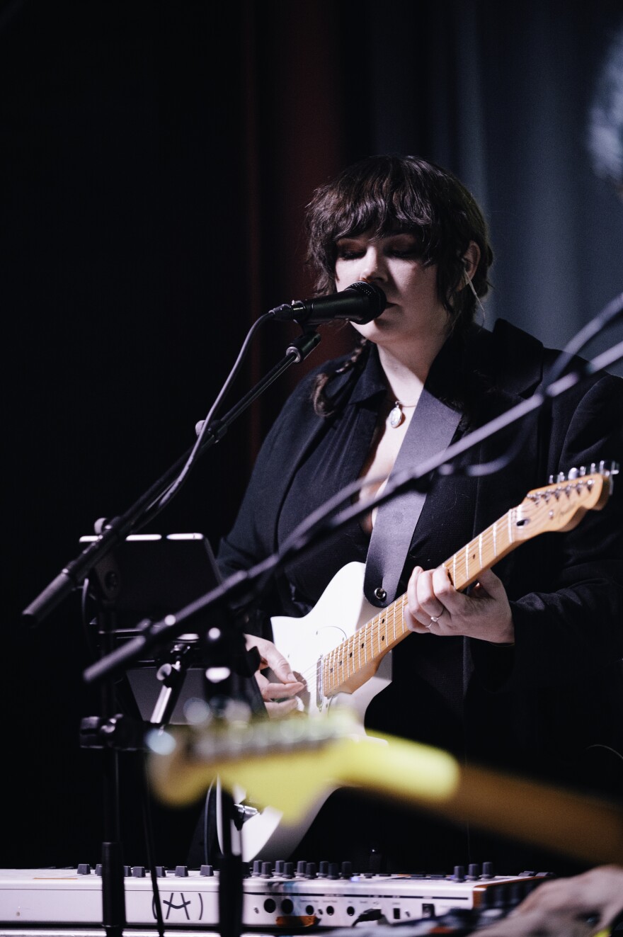 Marissa Kephart plays a white guitar and singing into a standing microphone.