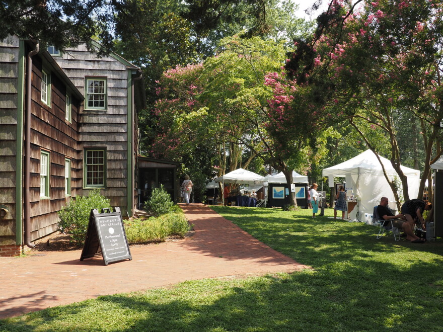 Image displaying tents of artists' work under the wooded canopy at Rehoboth Art League's wooded campus.