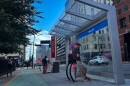 Two people lean against a curved structure under a bus shelter. Others stand near the bus stop.