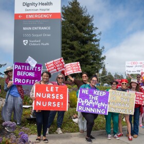 A group of people hold signs that say things like, "let nurses nurse," and "we keep the department running," stand on a street corner outside the hospital. 