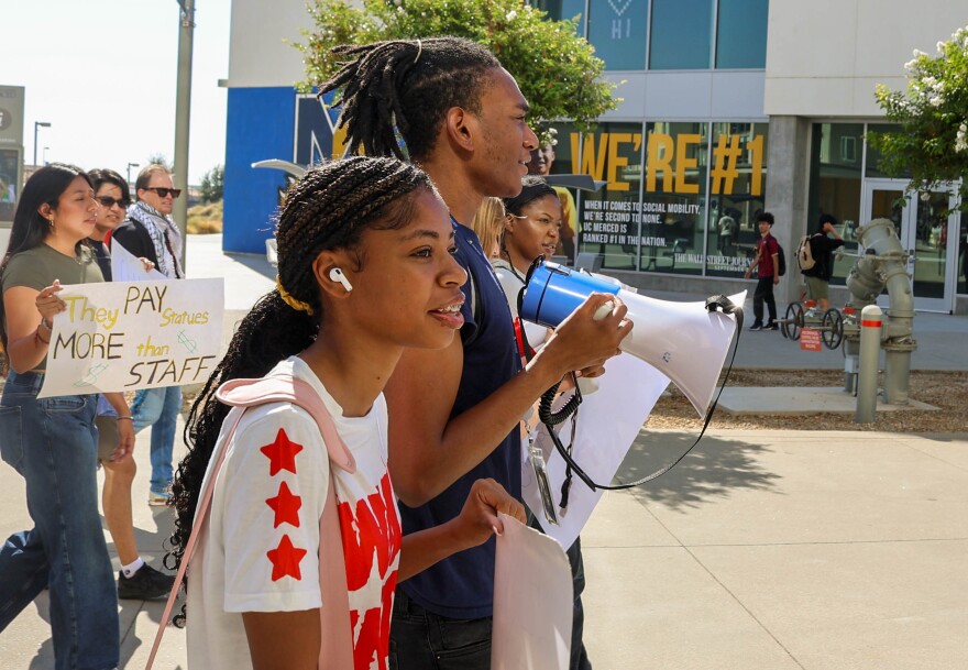 Brenden Wilson speaks into the megaphone as students march past a wall showing UC Merced’s recent Wall Street Journal No. 1 ranking in social mobility.