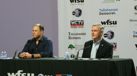 Two men sit at a table in front of microphones staring forward.