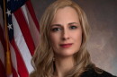 A woman with long blonde hair is wearing a black blazer over a red blouse and posing next to an American flag.