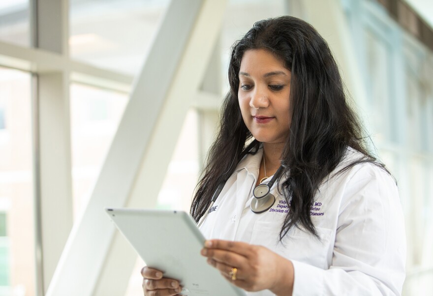 A woman with long hair, wearing a white coat and stethoscope, looks at a tablet.