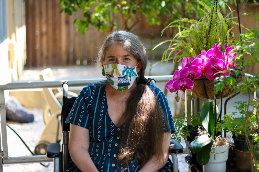 Ingrid Tischer. Ingrid is sitting in a chair on her front porch next to a bright magenta orchid. She is in her mid 50's and has gray and brown hair. She's wearing a white face mask with a plant pattern on it, and a patterned blue dress.