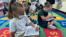 School children wear masks inside class while drawing on white boards. 