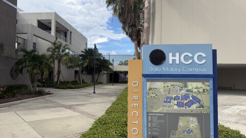 A blue and tan directory sign with a map of Hillsborough College's Dale Mabry campus stands in front of the entrance of grey classroom buildings and trees lining the walkway.