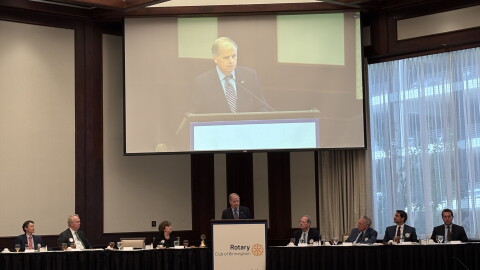 Doug Jones stands behind a podium with people on either side sitting along a table. A screen with his image is above him.