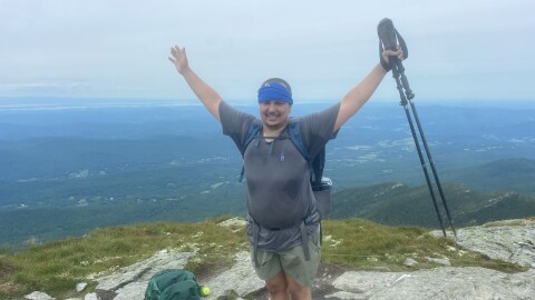 Sunshine Orta raises his hands in celebration, at the top of a hike, with a scenic vista behind him.
