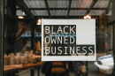 Black owned business sign taped on a window, seen from outside a store.