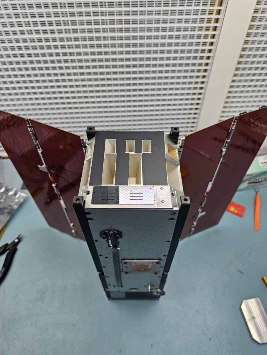 A small satellite on a lab bench. It has red solar panel wings and small compartments along its top.