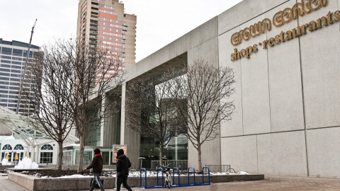 Outdoors photo showing the exterior of a large concrete building with trees in the front and two people walking in the foreground. A sign on the building reads: "Crown Center, shops and restaurants."