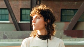A woman looks off camera with a flat expression while standing outside against the backdrop of an old industrial building.