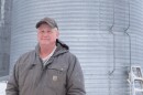 A man in a jacket and cap stands in front of a large metal grain bin