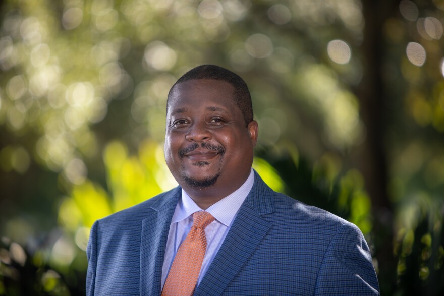 African-American man in blue suit and orange tie smiles at camera.