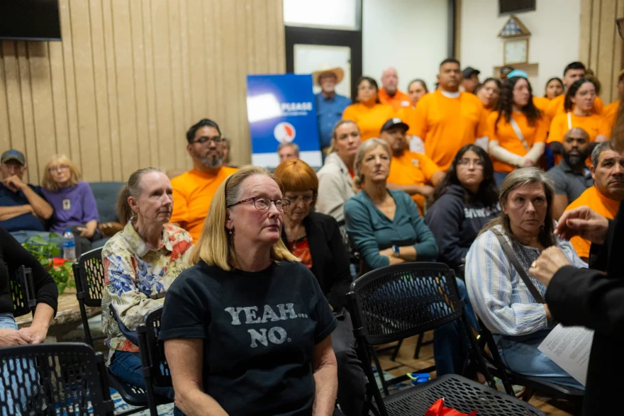 Residents sit and stand at San Marcos' City Council meeting, many are wearing matching orange shirts. A women in the front wears a dark shirt that reads 'Yeah... No.'