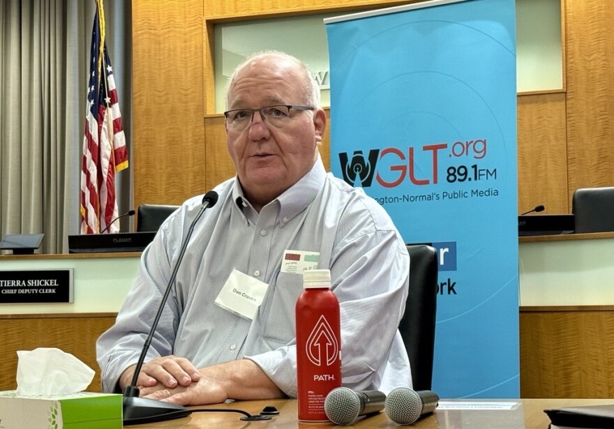A man in a button-down shirt sits at a desk with a microphone pointed at him. 