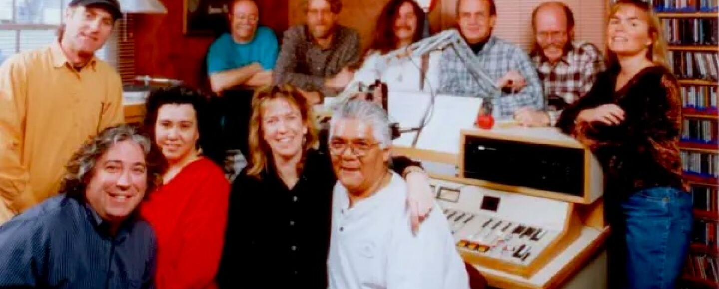 A group of 11 smiling people face the camera, they are seated and standing in a radio control room with a control board and a shelf of CDs in the back.
