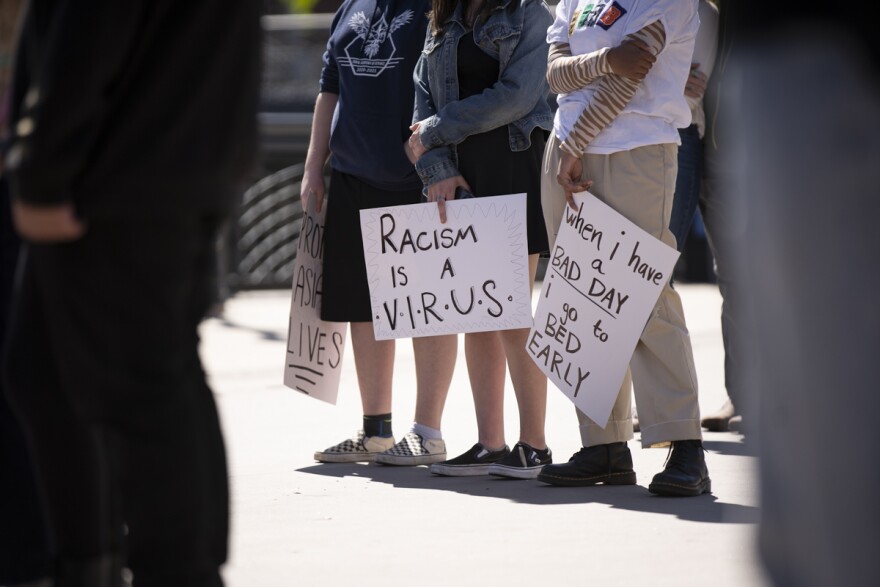 A photo outdoors of three protesters holding signs. Two signs that are legible say, “Racism is a virus,” and “When I have a bad day, I go to bed early.” The composition is framed by two out-of-focus figures on each side of the image.