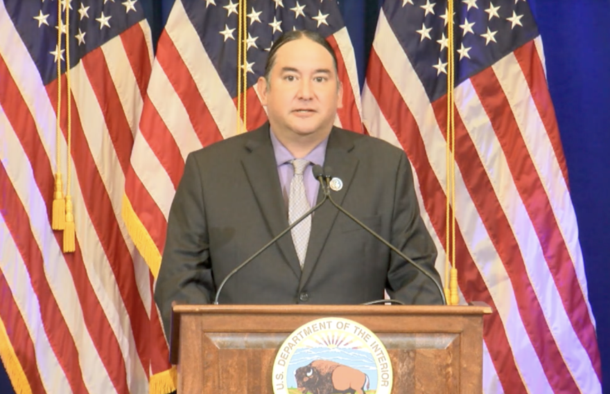 Hemene James stands behind a podium that has the Department of the Interior seal in front of it. He is wearing a dark gray suit and several American flags stand in the background.