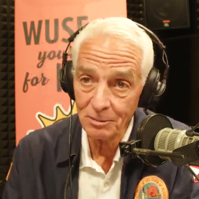 man with white hair is wearing headphones in a radio studio talking into a microphone. he is wearing black jacket over a white button shirt. the jacket has a round logo on it 