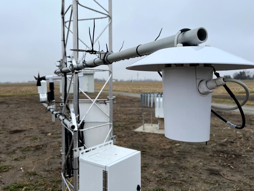 Weather monitoring gadgets are attached to a metal framework. The structure stands in the middle of a farm field.