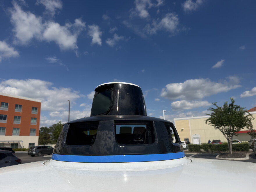 A black sensor sits on top of a white car that is sitting in a parking lot. There are multiple cameras seen within the bottom sensor.