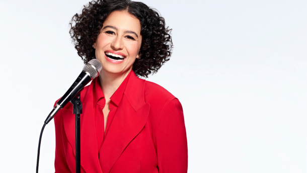 Headshot of Ilana Glazer, a comedian with curly black shoulder length hair, smiling big with a microphone to the left of her body. She's wearing a bright red suit jacket with the same color red button up shirt underneath. 
