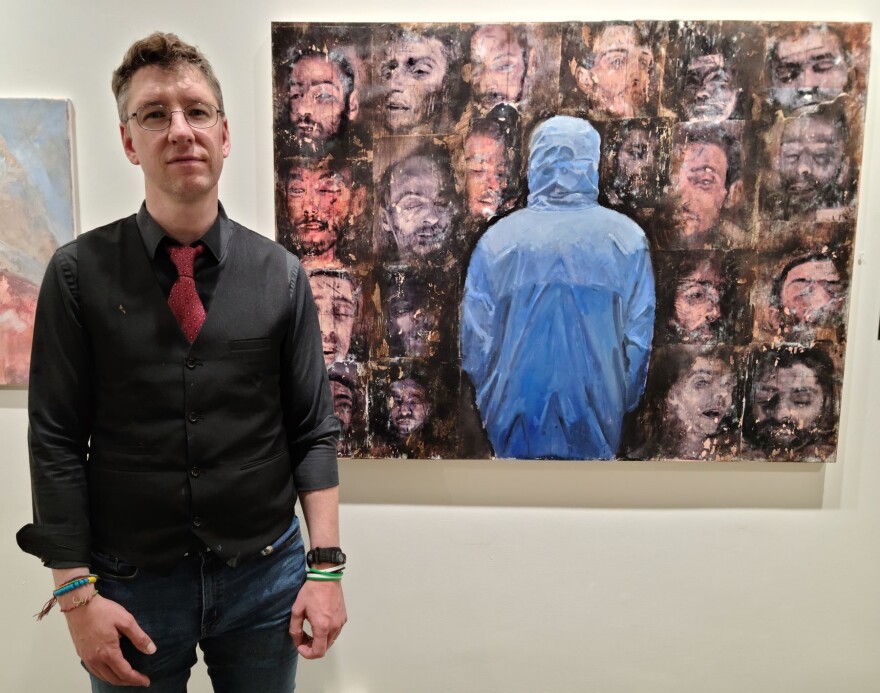 Marc Nelson next to his painting of Caesar (in the blue coat). “The images in the picture around him are drawings of faces that appeared in these thousands of photos that he snuck out of Syria.”