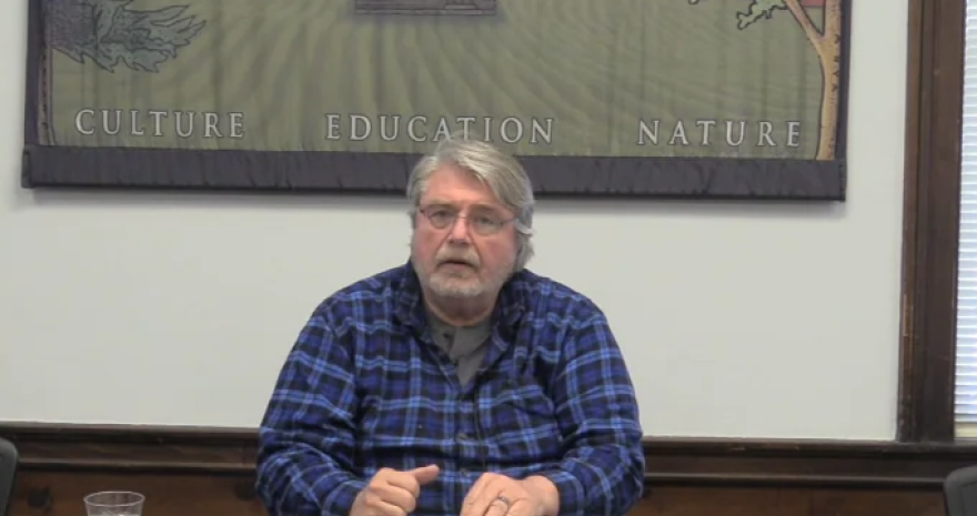 A white man with a beard and glasses in a blue flannel sits at a table