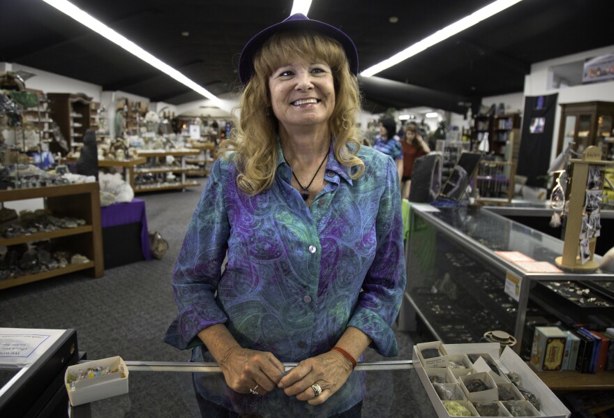 A woman is standing at a counter inside the store and smiling, but not looking at the camera. She is wearing a purple paisley shirt and purple hat. Behind her are tables and shelves baring crystals, rocks and minerals.