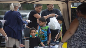 A family picks up bags of food from a canvas pop up tent in front of a school. 