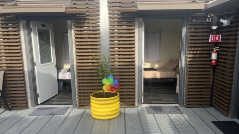 Two tiny home units with brown paneled exteriors have their doors open, showing beds inside. A yellow plant pot is outside between the two units.
