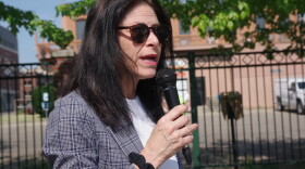  Michigan Attorney General Dana Nessel is wearing dark sunglasses, a white shirt and black and white plaid blazer at Arcadia Creek Festival Place before a Kalamazoo Pride event.  She's holding a microphone and has a black scrunchie hair tie around her wrist. 