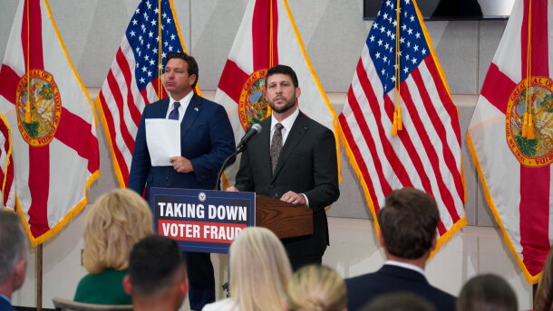 Two men in suit and ties by podium that reads "Taking Down Voter Fraud." A bunch of flags are behind them 