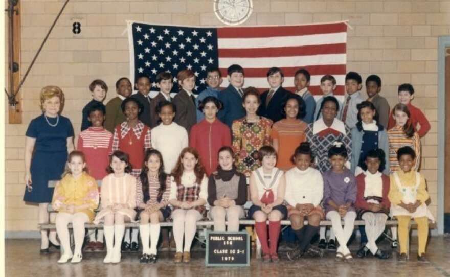 A class photo of fifth graders from the late 60s in front of the U.S. flag. There is an even split of Black and white students, mostly grouped in clumps. In the front row, four girls have their legs crossed.